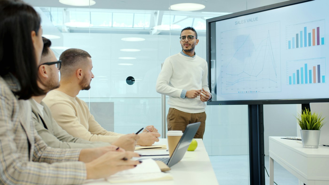Arabian man employee is giving presentation to group of colleagues in conference room speaking and showing infographics on digital whiteboard.